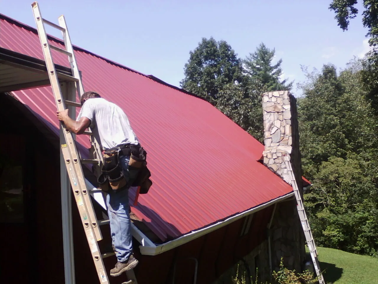 Roofer on a ladder beside a red metal roof and stone chimney — Soffit & Fascia Repair by Roofing Brothers in Mcchord Afb
